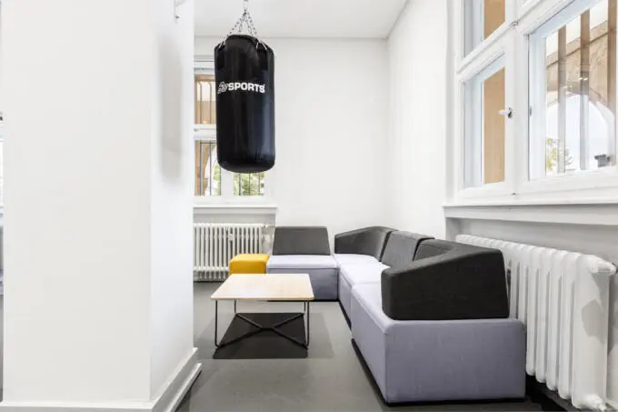 Heller Büroraum mit schwarzem Boxsack, grauer Eckcouch und kleinem Holztisch vor großen weißen Fenstern