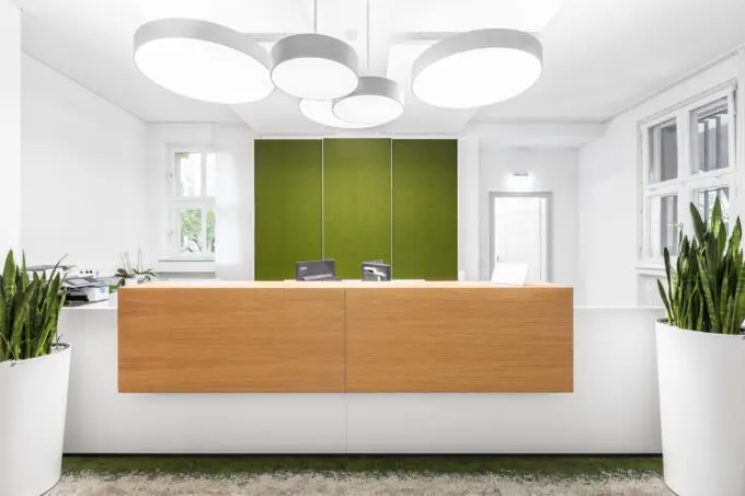 Frontale Ansicht eines modernen Empfangstresens mit Holzfront, runden Deckenleuchten und grünen Wandpaneelen in einem hellen Raum