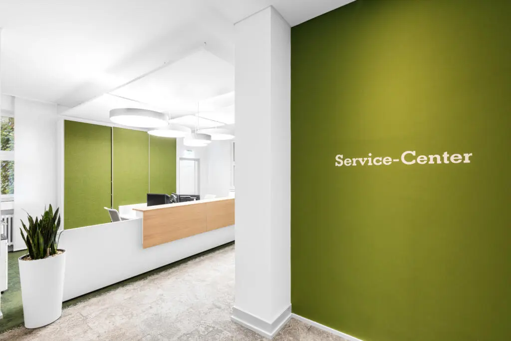 Innenansicht eines modernen Service-Centers mit grüner Wand, weißem Schriftzug und Empfangstresen mit Holzfront
