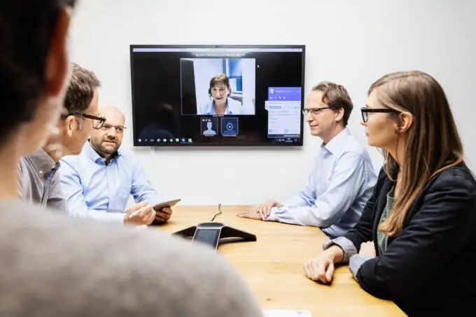 Vier Personen sitzen an einem Holztisch in einem hellen Besprechungsraum und führen eine Videokonferenz mit einer Teilnehmerin auf einem großen Wandbildschirm.