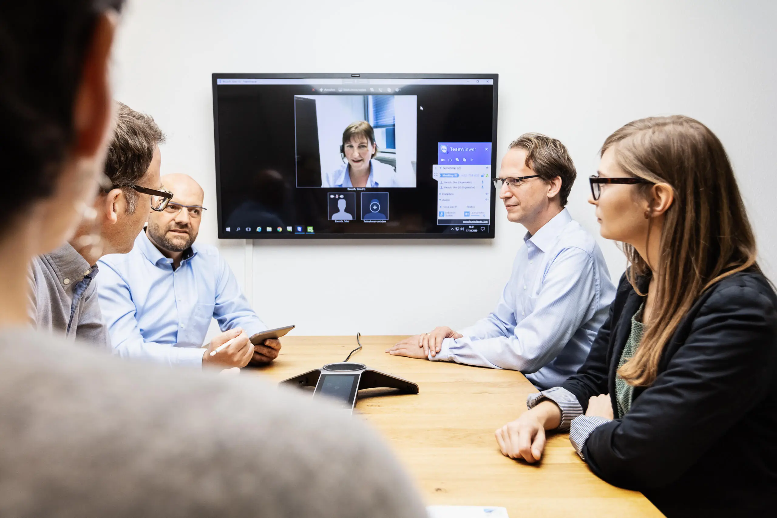 Vier Personen sitzen an einem Holztisch in einem hellen Besprechungsraum und führen eine Videokonferenz mit einer Teilnehmerin auf einem großen Wandbildschirm.