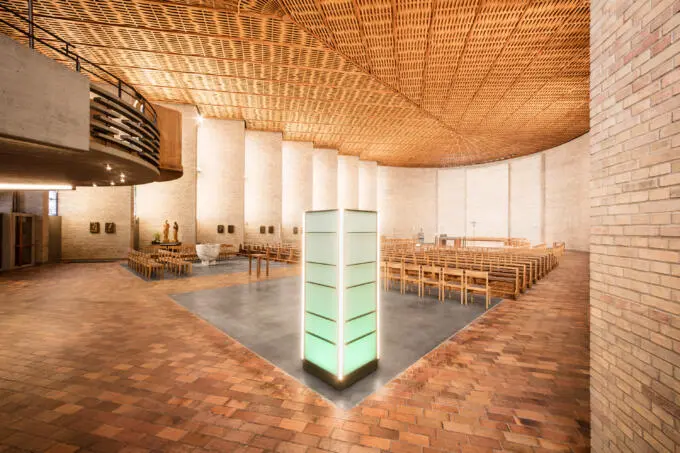 Innenraum einer modernen Kirche mit radialer Holzdecke, Backsteinwänden und zentraler Lichtsäule