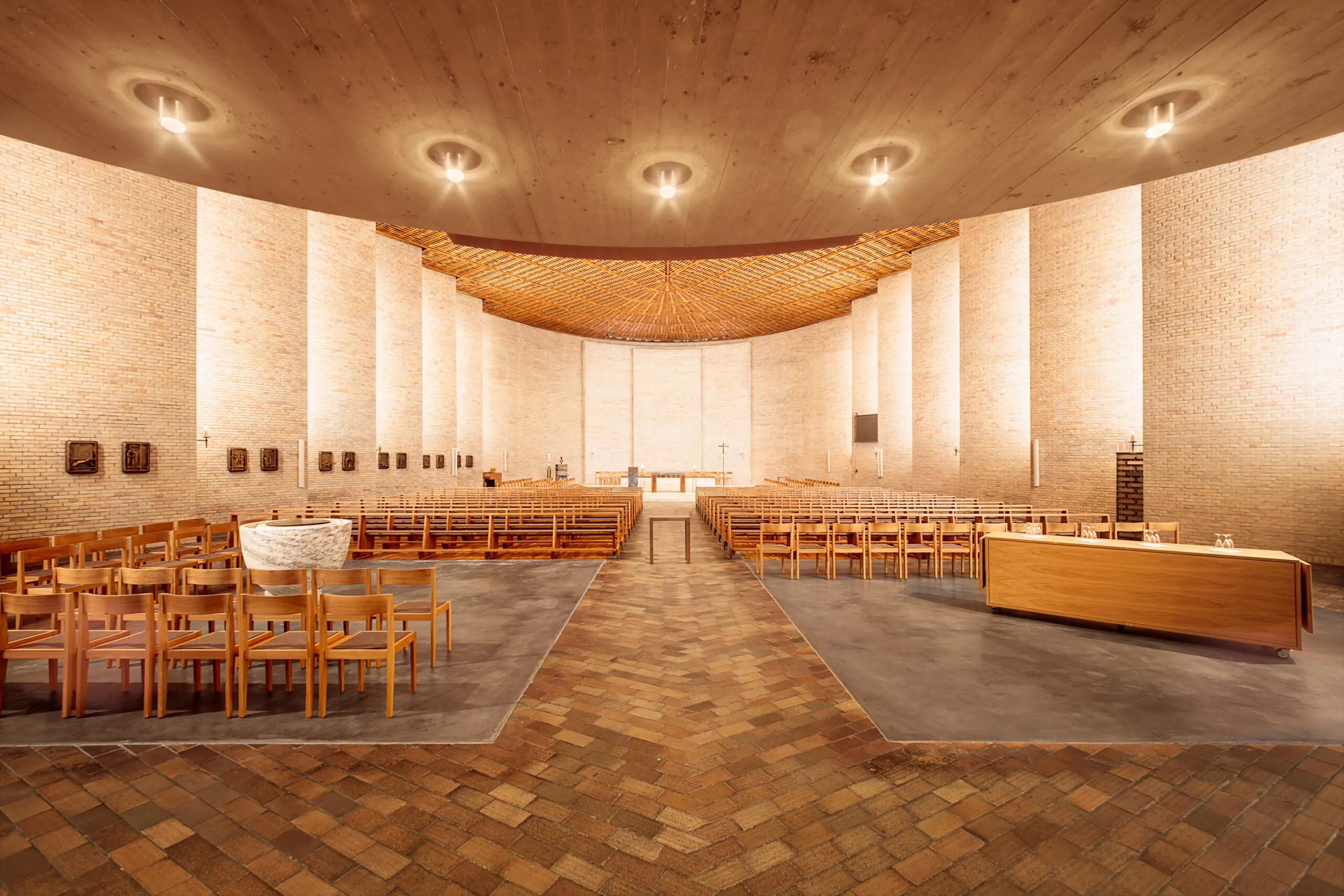Innenansicht einer modernen Kirche mit geschwungenen Backsteinwänden, Betondecke mit runden Leuchten und hölzernen Kirchenbänken