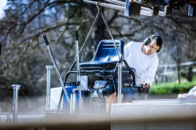 Bauarbeiter mit Gehörschutz prüft eine an Hebegurten hängende Aufsitz-Betonglättmaschine auf einer Baustelle