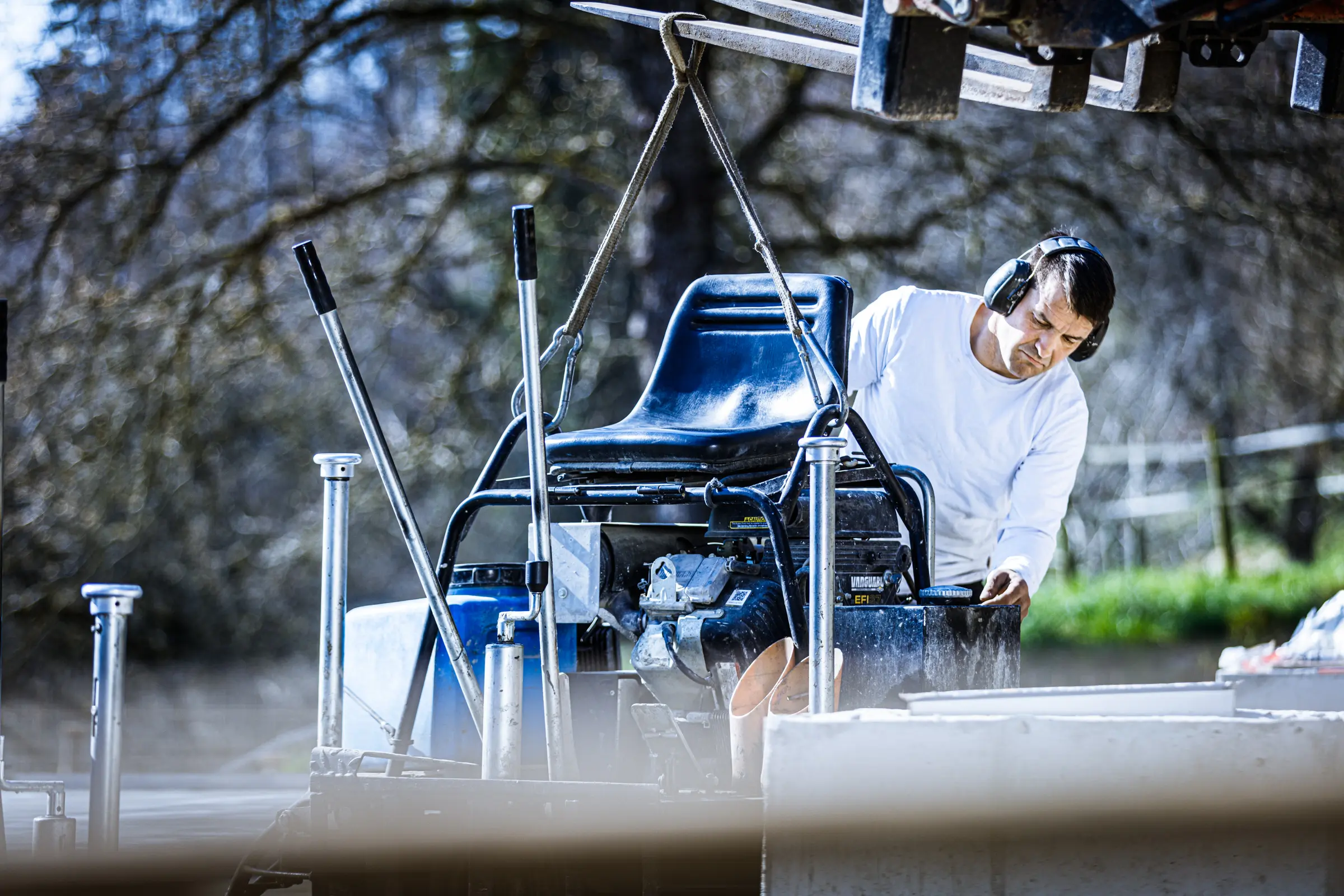 Bauarbeiter mit Gehörschutz prüft eine an Hebegurten hängende Aufsitz-Betonglättmaschine auf einer Baustelle