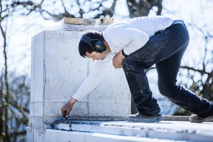 Bauarbeiter mit Gehörschutz bearbeitet kniend eine Betonoberfläche auf einer Baustelle im Freien