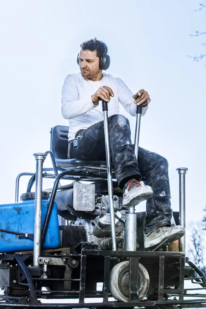 Bauarbeiter mit Gehörschutz bedient einen Aufsitz-Flügelglätter vor hellem Himmel