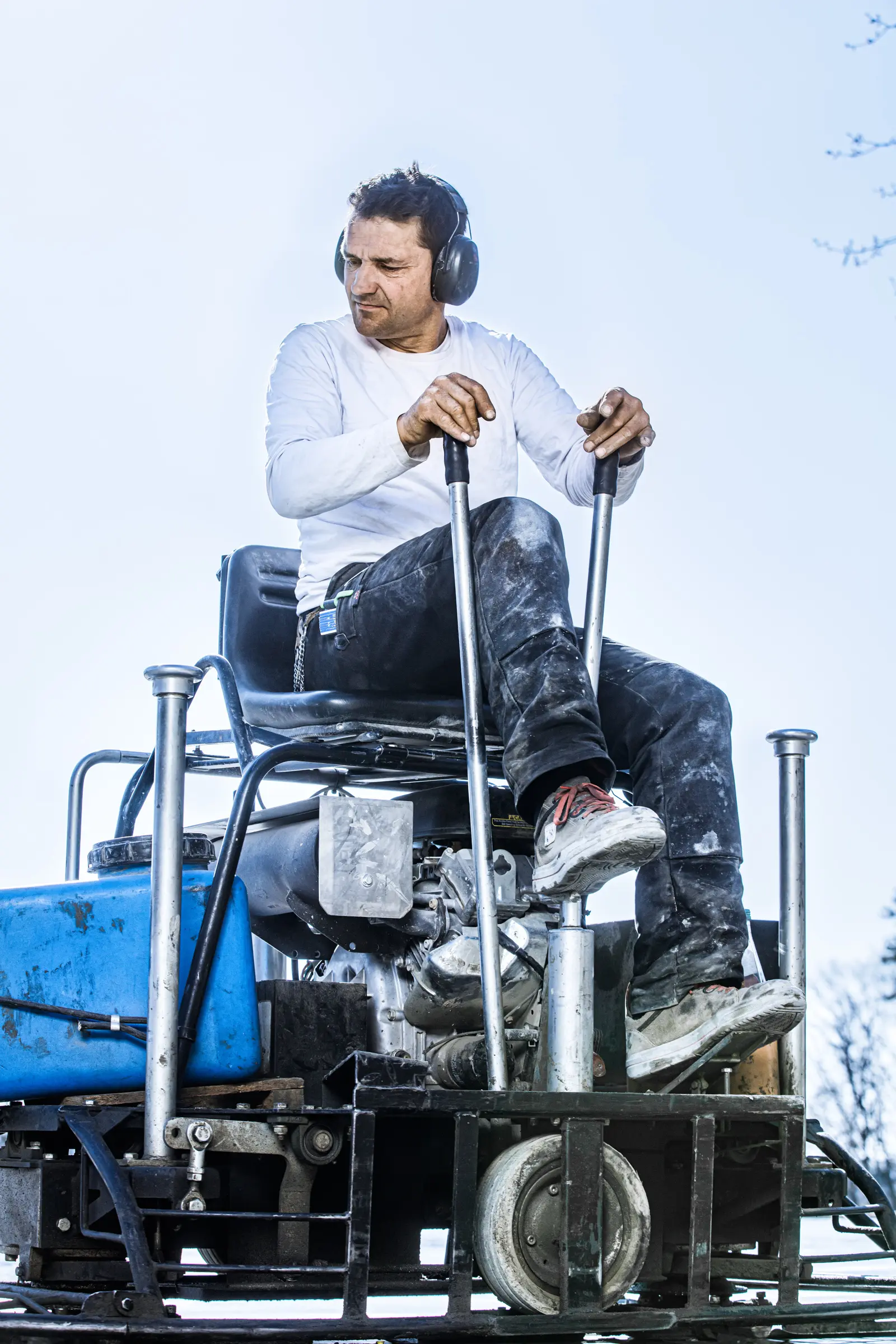 Bauarbeiter mit Gehörschutz bedient einen Aufsitz-Flügelglätter vor hellem Himmel