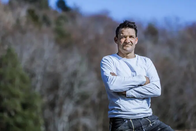 Porträt eines Handwerkers in weißem Shirt und Zunfthose mit verschränkten Armen vor unscharfem Naturhintergrund