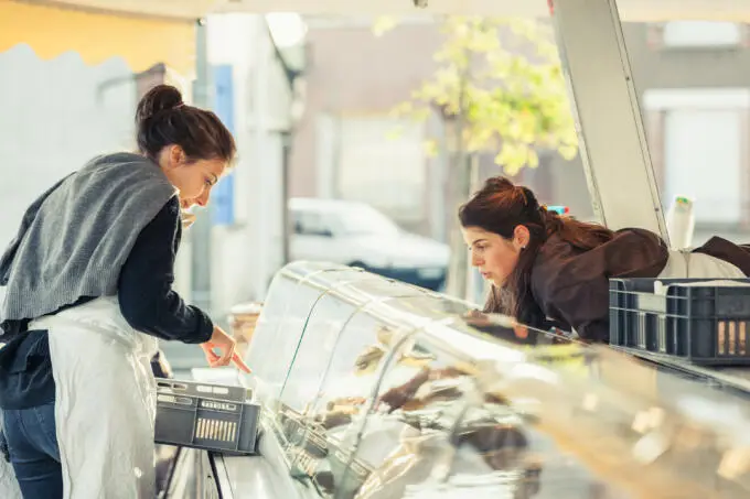 Eine Frau mit Schürze zeigt auf Waren in einer Glasvitrine und spricht mit einer Kollegin an einem Marktstand.