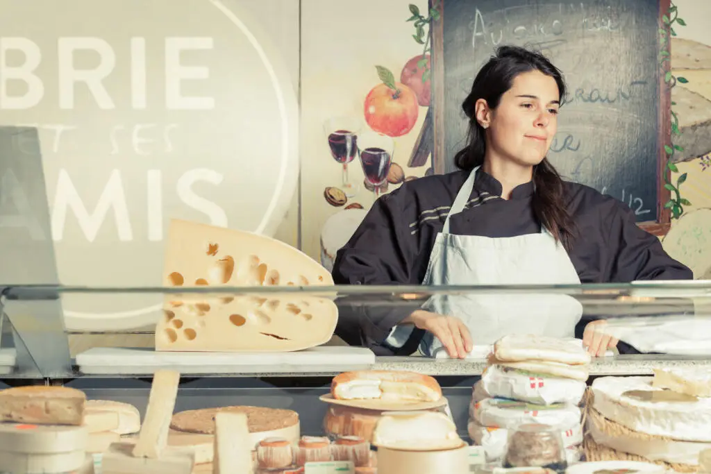 Eine Verkäuferin in Schürze steht hinter einer gut gefüllten Käsetheke mit verschiedenen Käsesorten und einem großen Stück Hartkäse im Vordergrund.