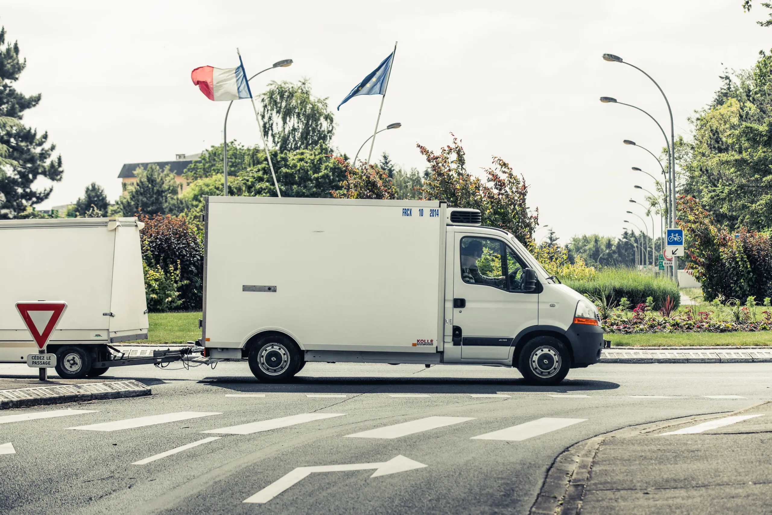 Ein weißer Kühltransporter mit passendem Anhänger fährt bei Tageslicht durch einen Kreisverkehr mit sichtbaren Straßenmarkierungen und Flaggen im Hintergrund.