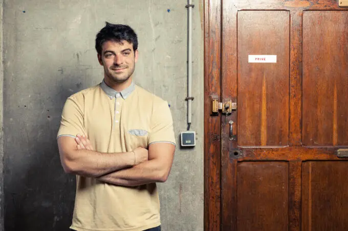 Ein junger Mann steht mit verschränkten Armen lächelnd vor einer grauen Betonwand und einer dunklen Holztür mit der Aufschrift Privé.