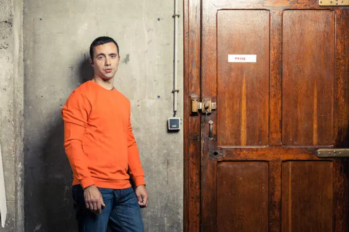 Ein Mann in einem orangen Pullover steht vor einer grauen Betonwand neben einer dunklen Holztür mit einem Privé-Schild.