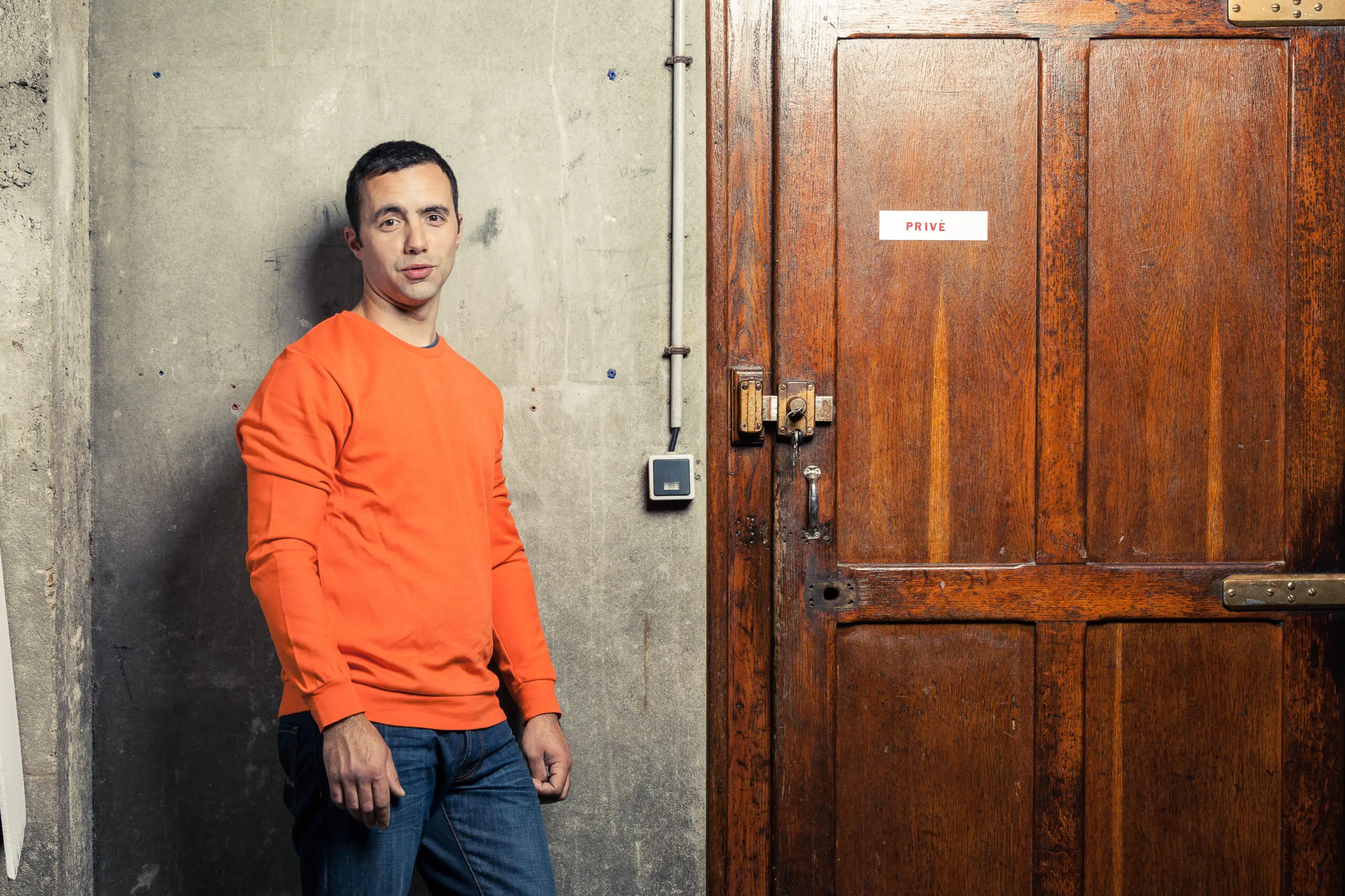 Ein Mann in einem orangen Pullover steht vor einer grauen Betonwand neben einer dunklen Holztür mit einem Privé-Schild.