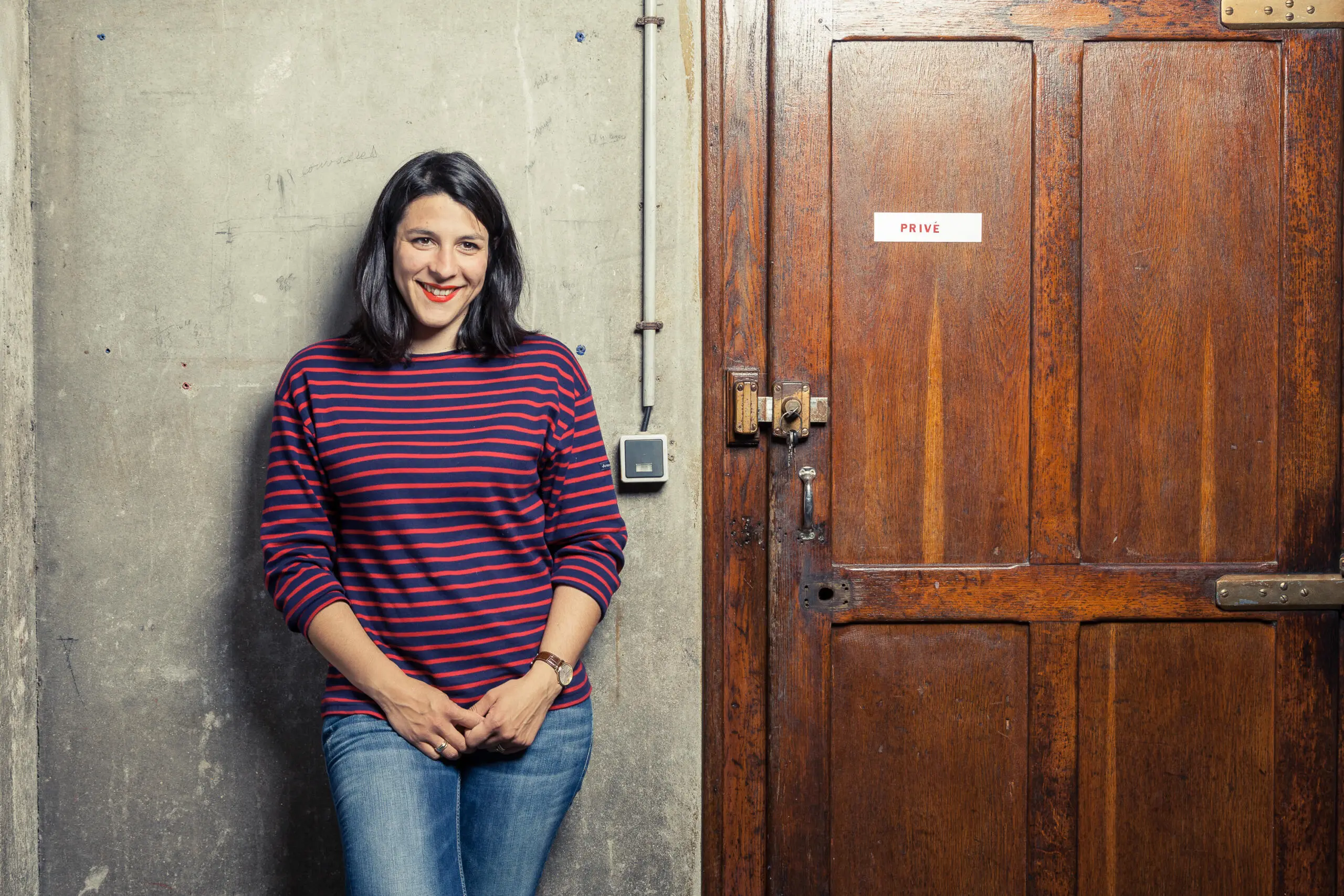 Eine lächelnde Frau in gestreiftem Oberteil steht vor einer grauen Betonwand neben einer dunklen Holztür mit dem Schild Privé.