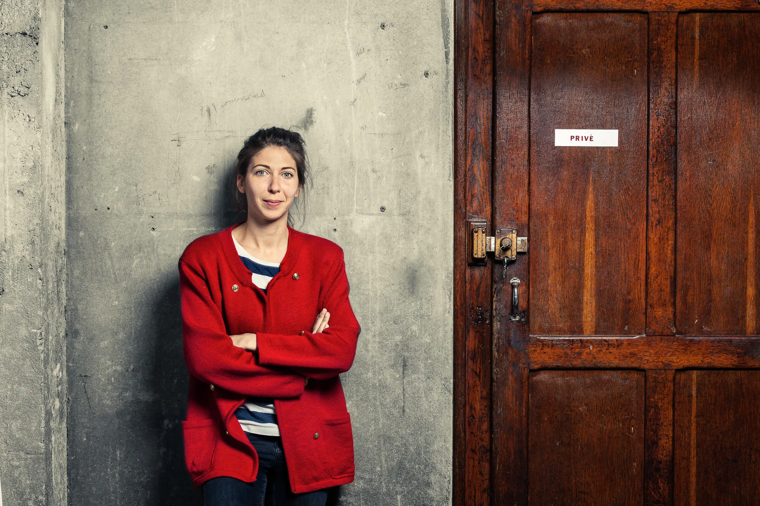 Eine Frau in roter Strickjacke steht mit verschränkten Armen vor einer grauen Betonwand neben einer dunklen Holztür mit der Aufschrift Privé.