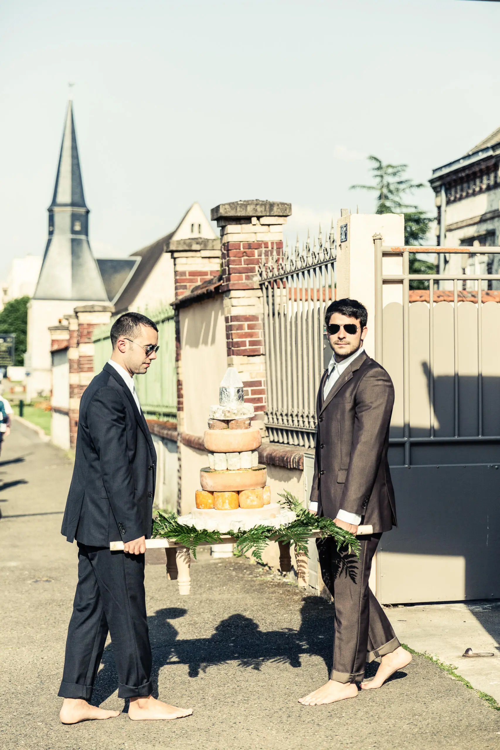Zwei barfüßige Männer in Anzügen tragen eine mehrstöckige Torte aus Käselaiben auf einer Holztrage im Freien.