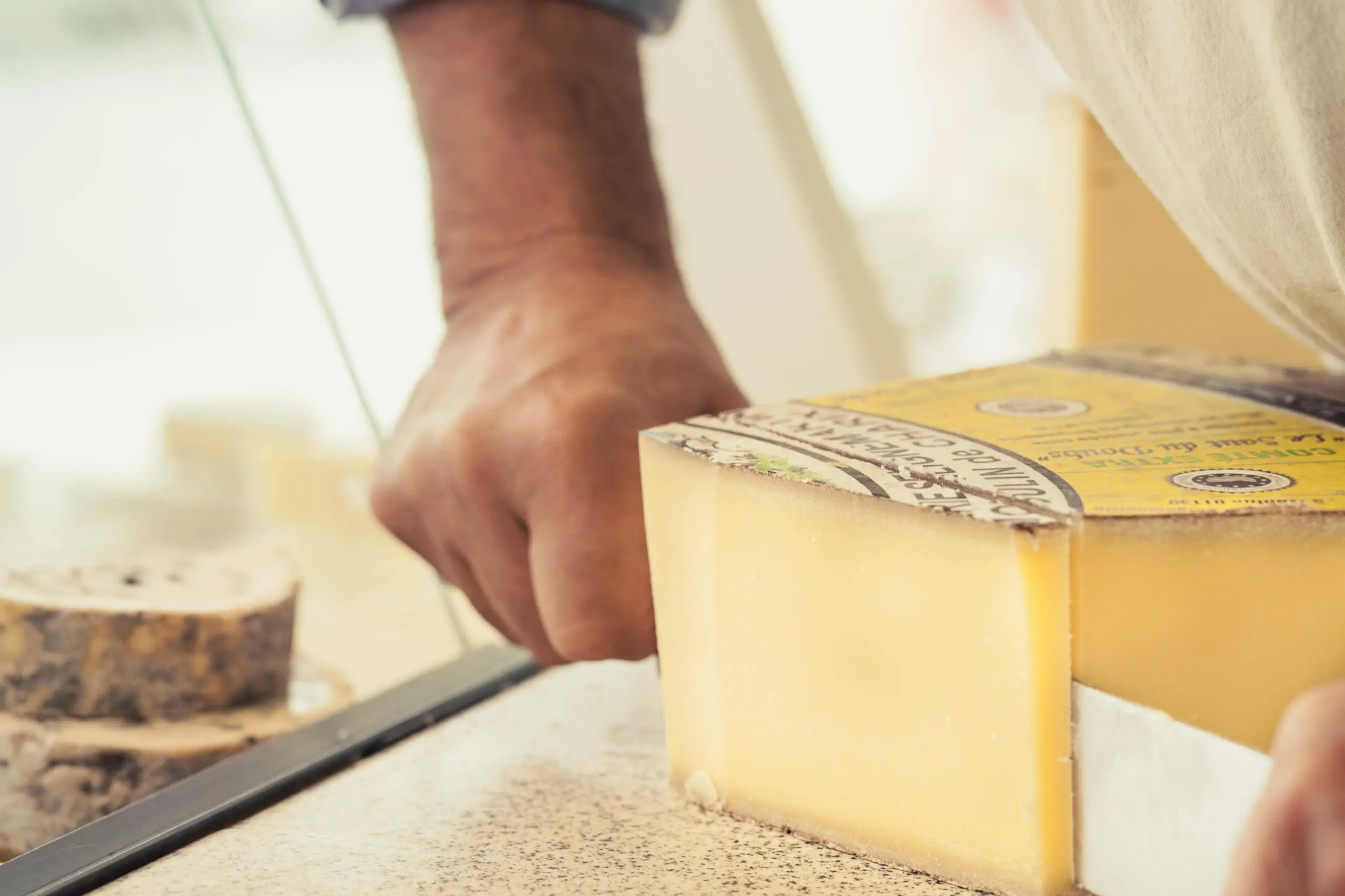 Eine Hand schneidet mit einem Werkzeug ein großes Stück Comté-Käse auf einer Arbeitsfläche an