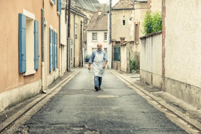 Ein Mann mit weißer Schürze und kariertem Hemd geht mittig auf einer schmalen Dorfstraße auf die Kamera zu.