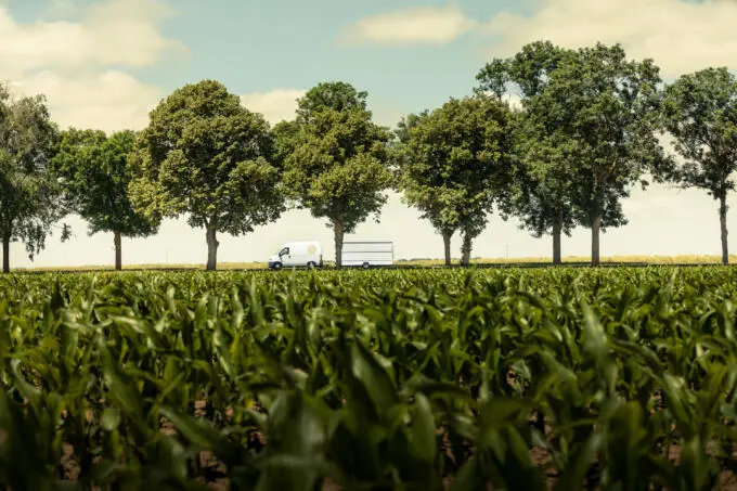 Ein weißer Lieferwagen mit Anhänger fährt auf einer von Bäumen gesäumten Landstraße, gesehen durch ein grünes Maisfeld unter blauem Himmel.