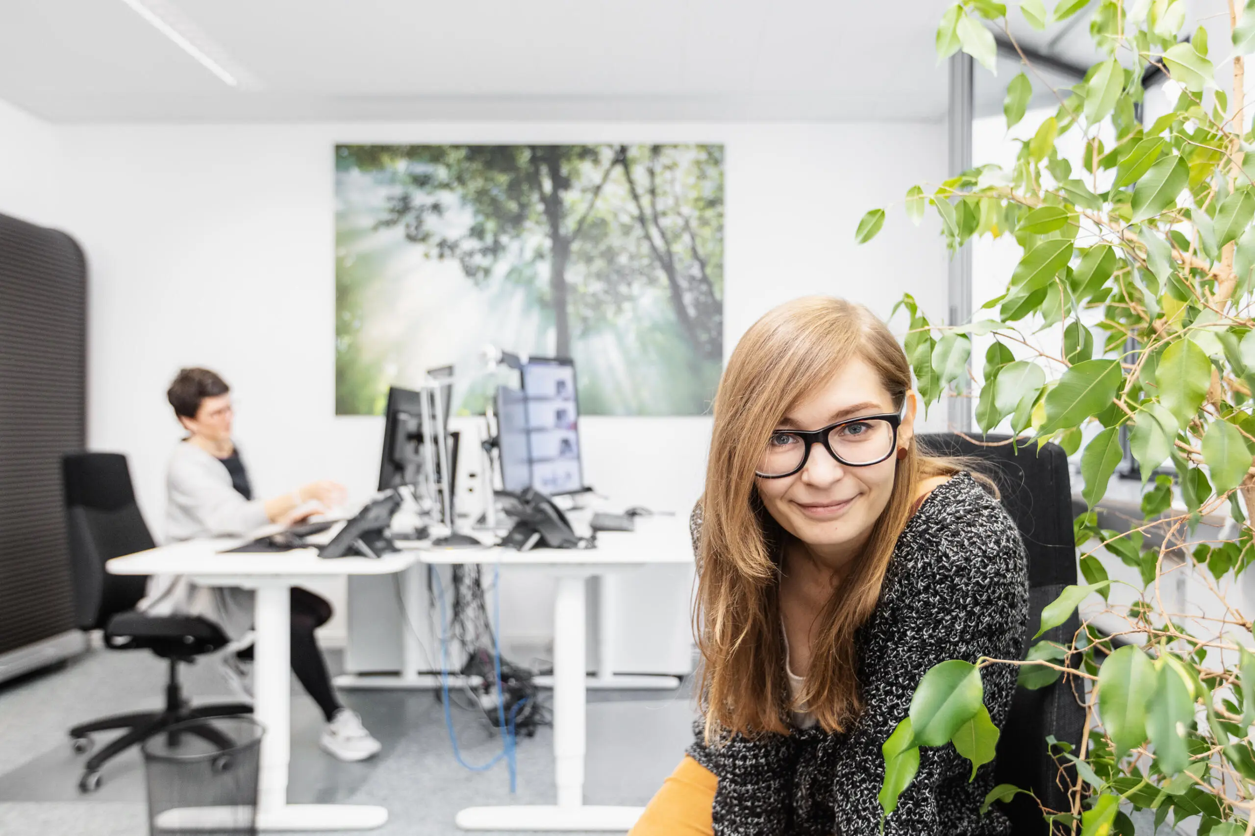 Eine lächelnde Mitarbeiterin mit Brille sitzt im Vordergrund eines hellen Büros neben einer Grünpflanze, während im Hintergrund eine Kollegin am Schreibtisch arbeitet.