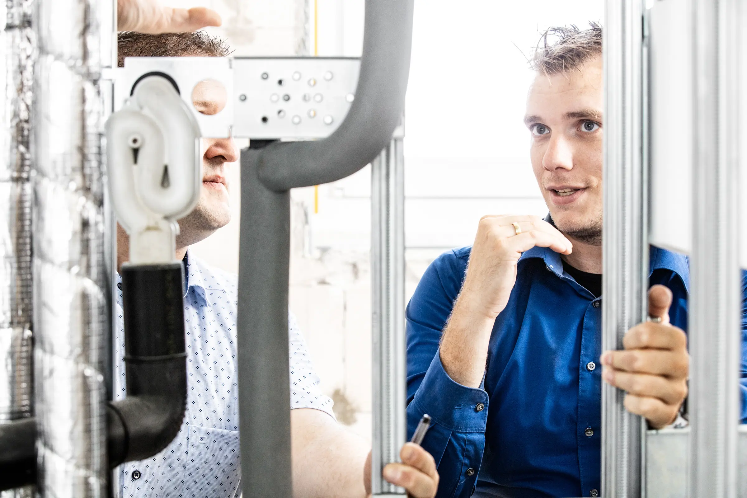Zwei Handwerker in blauen Hemden besprechen Sanitärinstallation zwischen Metallprofilen und isolierten Rohren auf einer Baustelle