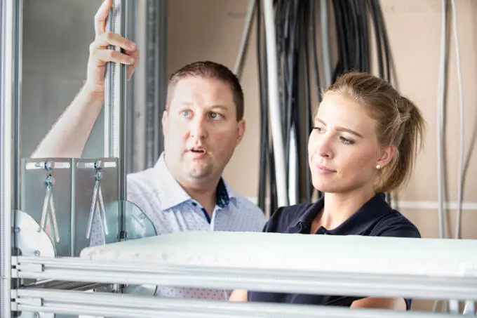 Ein Mann und eine Frau betrachten konzentriert eine Metallrahmenkonstruktion für Sanitärinstallationen vor einer Trockenbauwand mit Kabeln