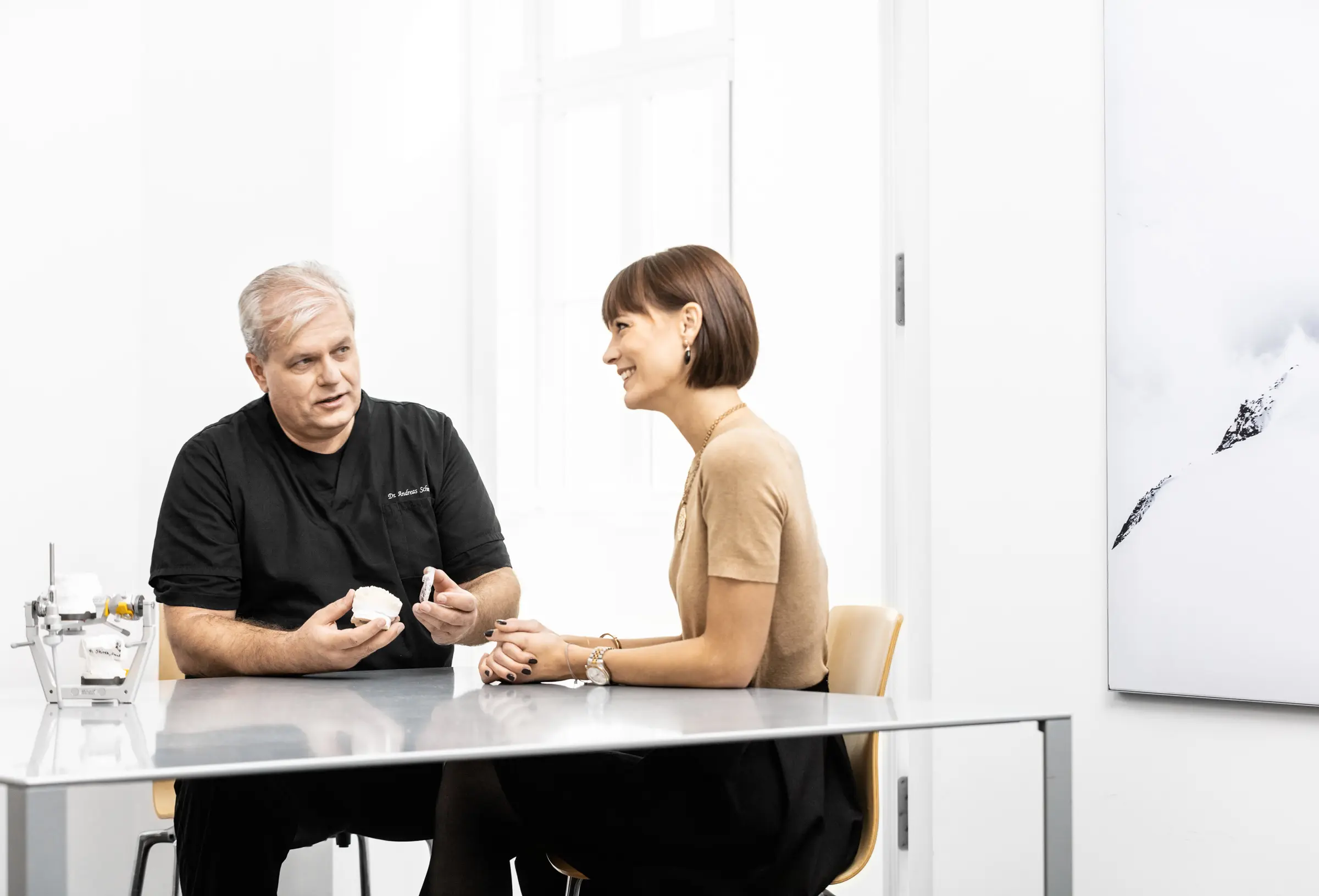 Zahnarzt in schwarzer Kleidung erklärt Patientin an einem Metalltisch ein weißes Kiefermodell in heller Praxisumgebung