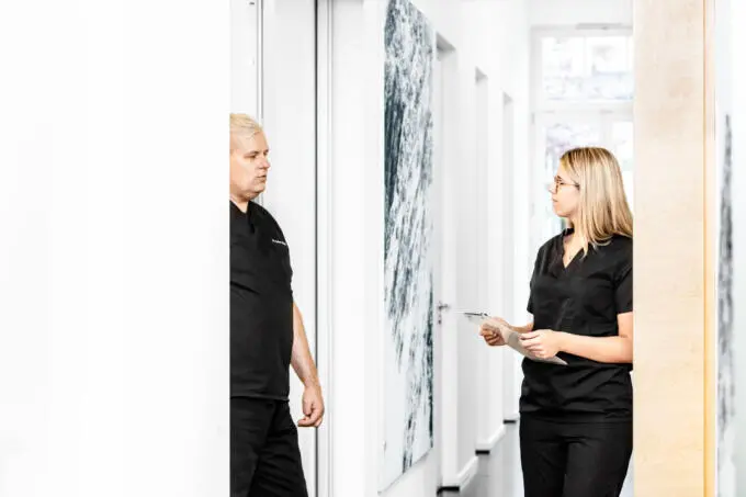 Zwei medizinische Fachkräfte in schwarzer Berufskleidung im Gespräch in einem hellen Praxisflur mit Wandkunst