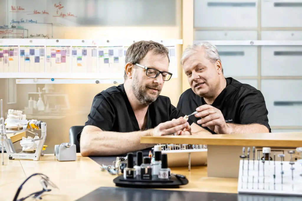 Zwei Zahntechniker in schwarzer Arbeitskleidung begutachten gemeinsam ein Werkstück am Labortisch mit Artikulator und Werkzeugen
