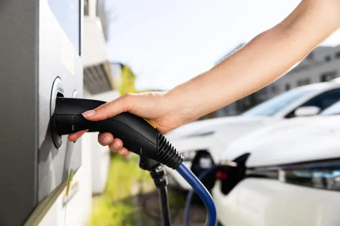 Nahaufnahme einer Hand, die einen Ladestecker in eine Ladestation für Elektroautos steckt, mit unscharfen Fahrzeugen im Hintergrund