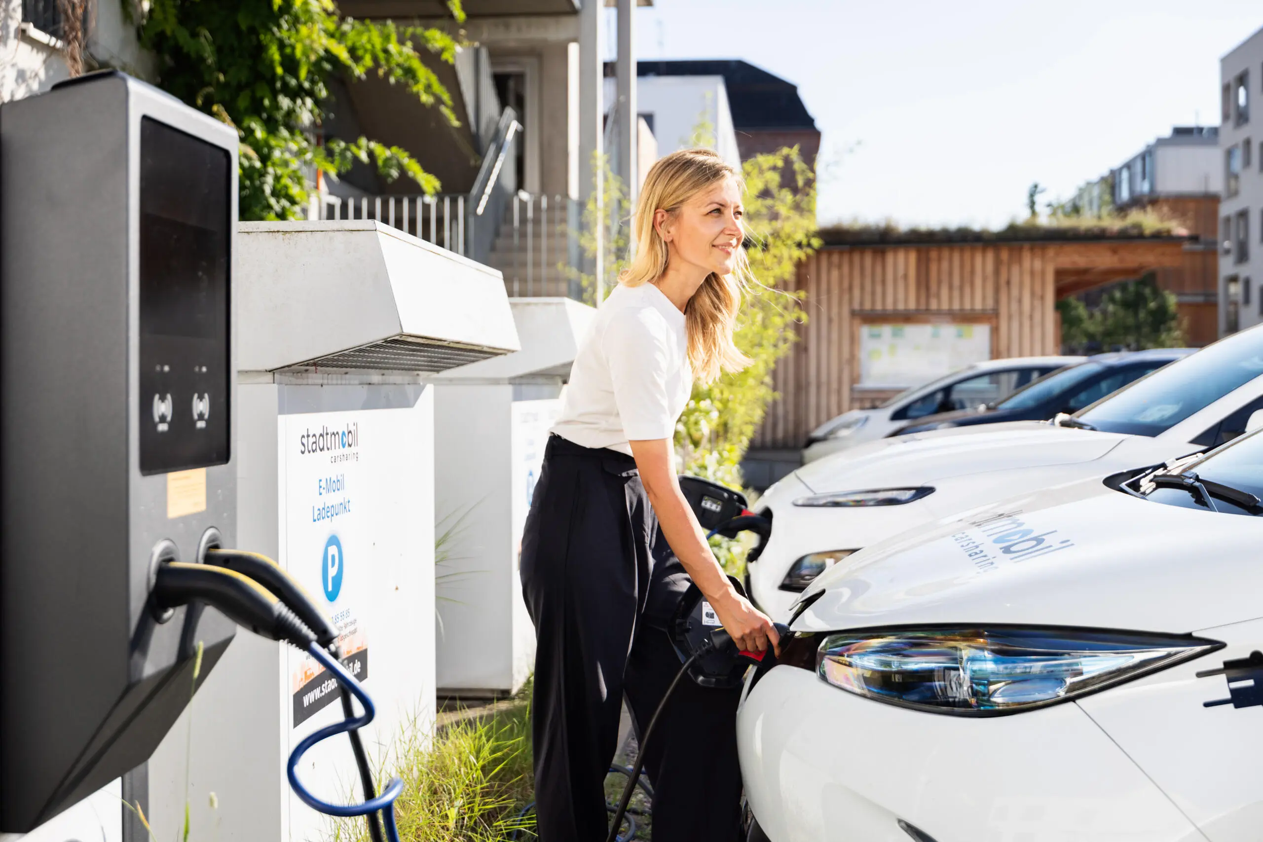 Frau schließt Ladekabel an weißes Elektroauto an einer Ladesäule im städtischen Außenbereich an