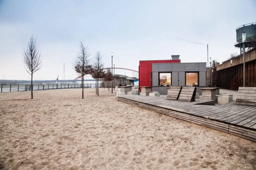 An outdoor sandy area features a wooden terrace with seating furniture and a modular kiosk building against an urban river backdrop.
