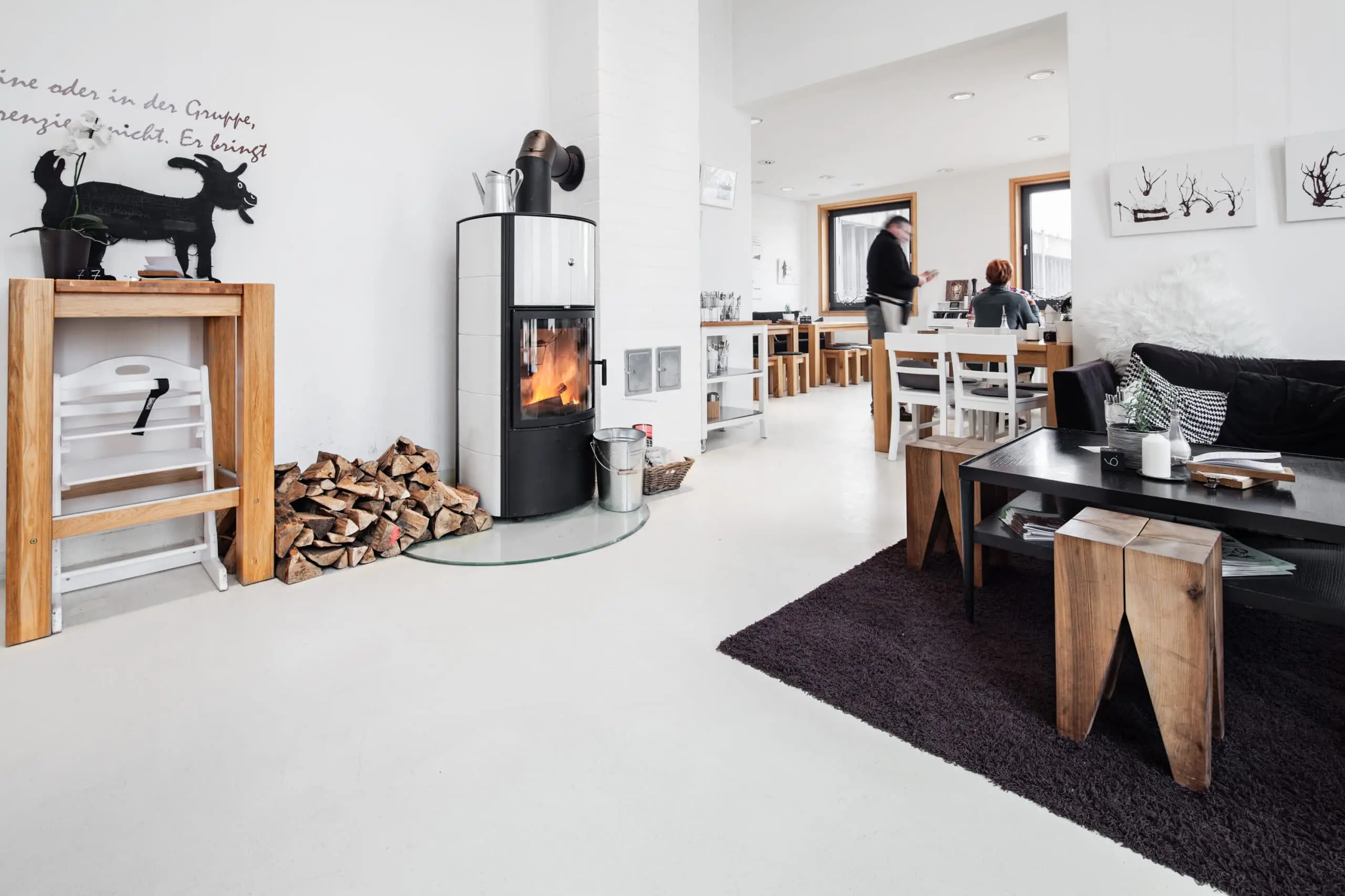 Heller Gastraum mit weißem Kaminofen, gestapeltem Brennholz und einer Sitzecke mit Holzhockern vor einem Essbereich.