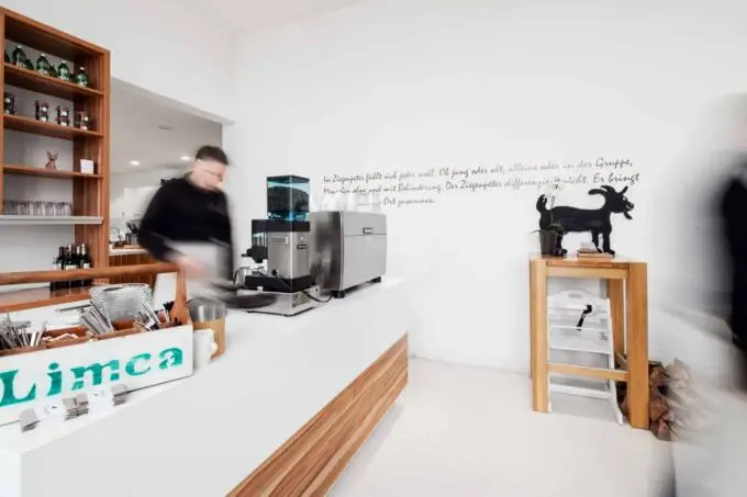 Helle Theke in einem Restaurant mit Kaffeemaschine und einer Servicekraft in Bewegungsunschärfe vor einer weißen Wand mit Text.