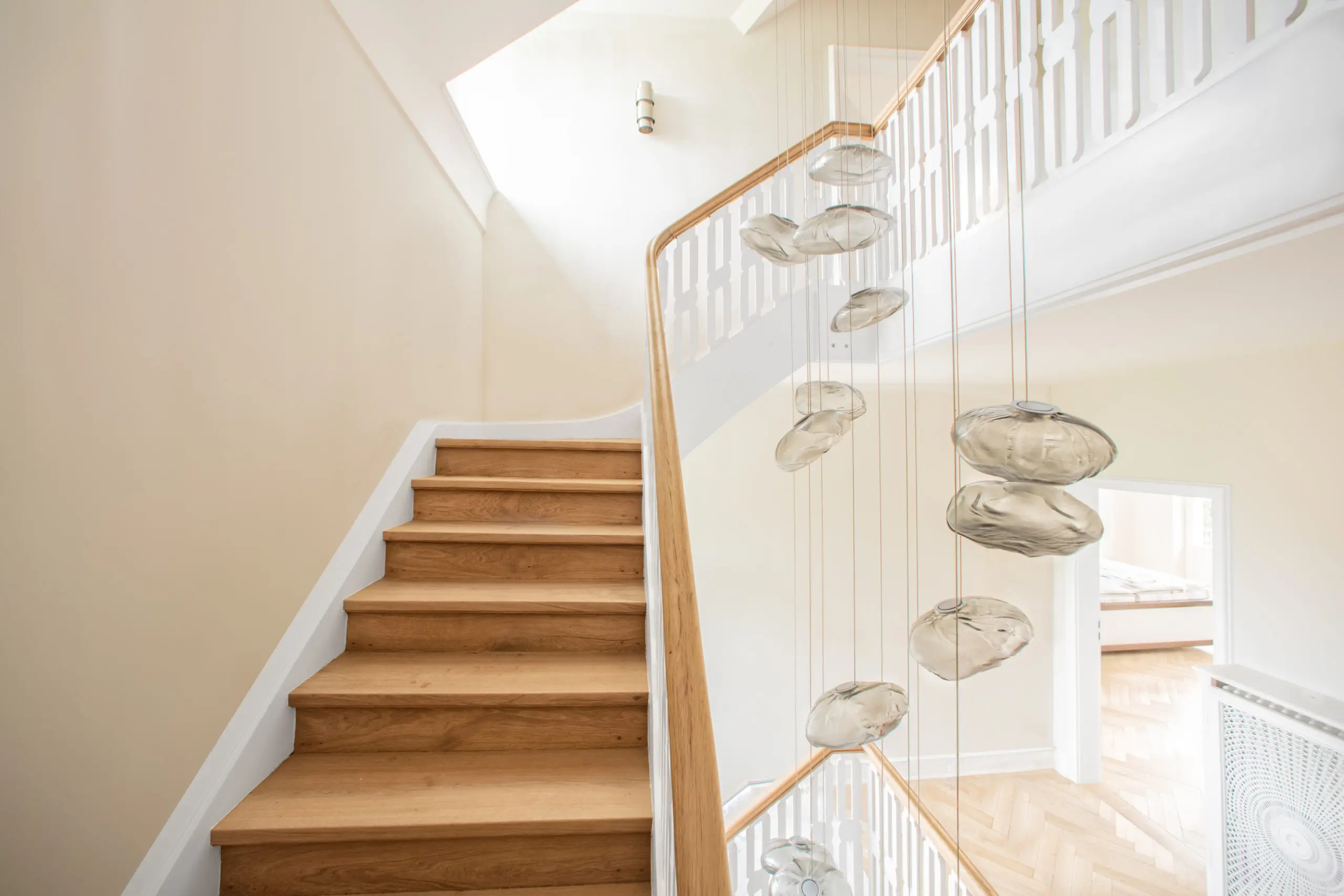 Helle Holztreppe mit weißem Geländer und moderner, mehrstufiger Glas-Pendelleuchte in einem lichtdurchfluteten Treppenhaus mit Fischgrätparkett.