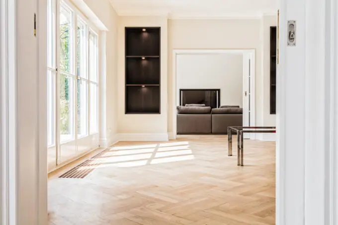 Ein heller Raum mit hellem Fischgrätparkett, bodentiefen Fenstern und einem dunklen Einbauregal, gesehen durch einen weißen Türrahmen.