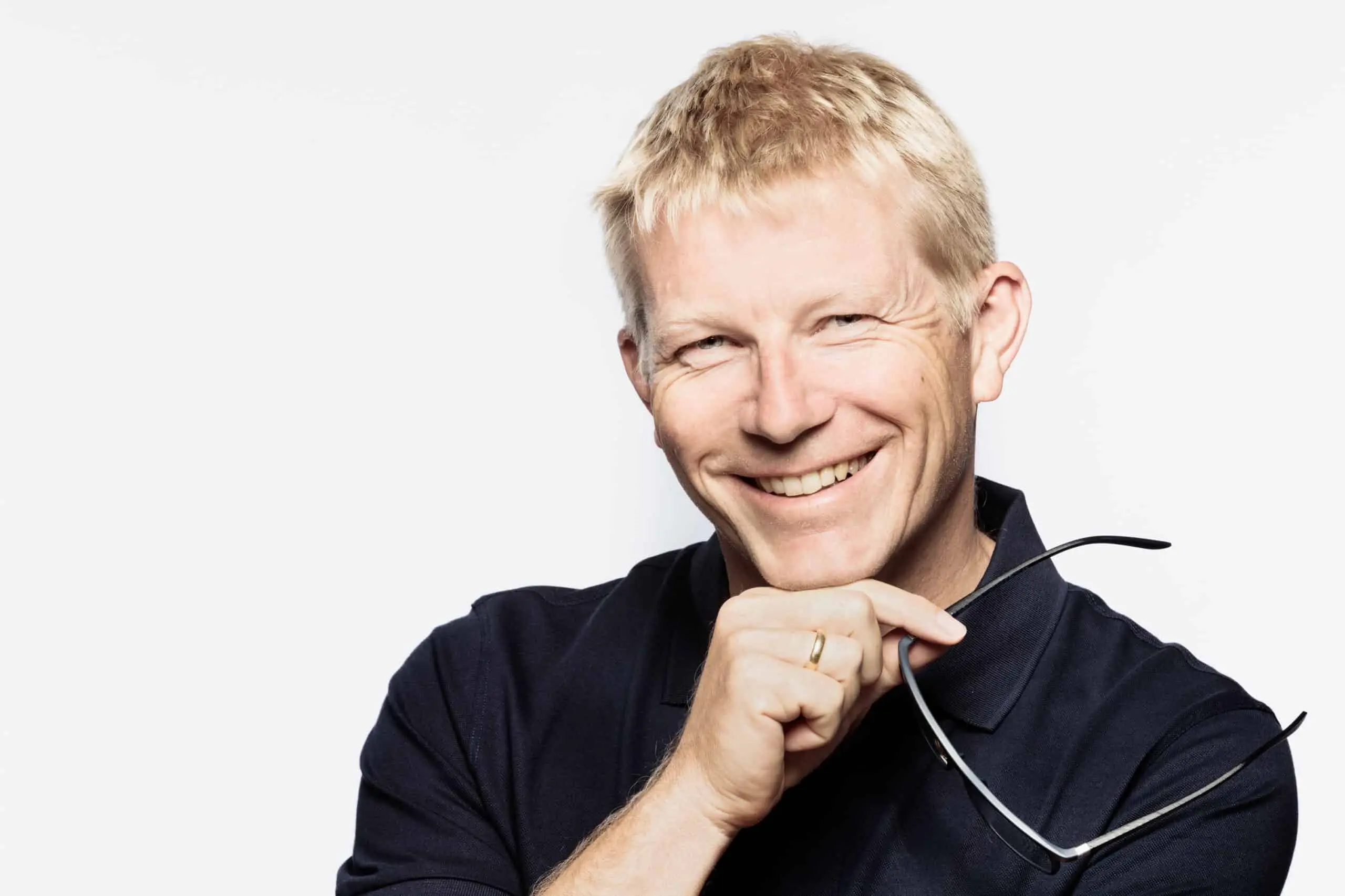 A smiling man with short blond hair holds a pair of glasses in his hand while wearing a dark blue polo shirt against a white background.