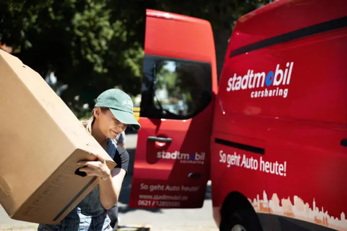 Frau trägt einen Umzugskarton neben einem roten Carsharing-Transporter mit offener Seitentür im Freien