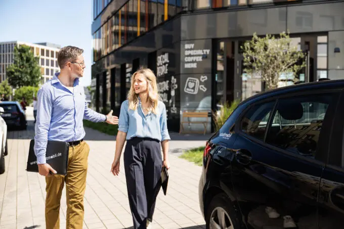 Mann und Frau in Business-Kleidung gehen an einem Carsharing-Auto vor modernem Bürogebäude vorbei