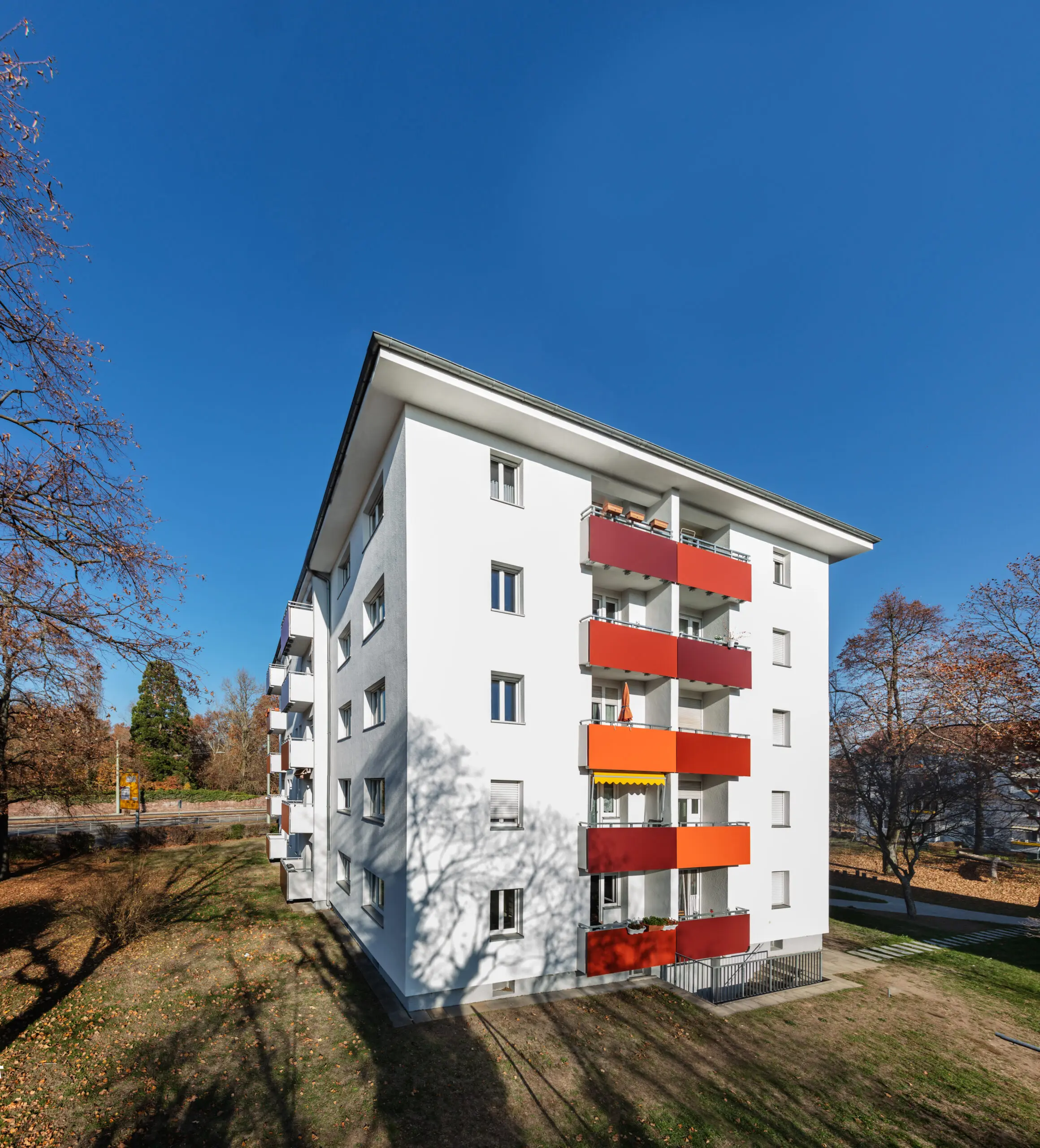 Außenansicht eines sanierten weißen Mehrfamilienhauses mit roten und orangen Balkonen unter blauem Himmel im Herbst