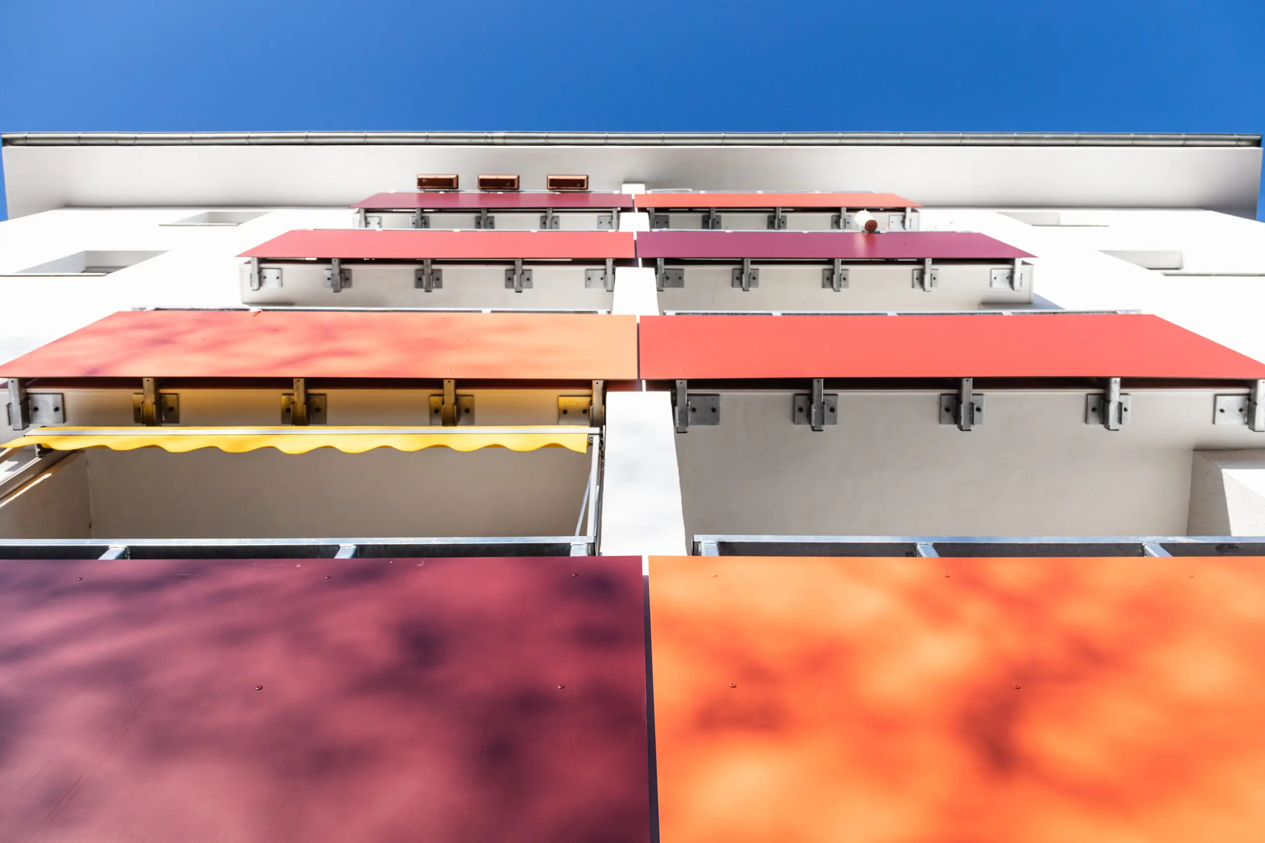 Steile Untersicht einer weißen Fassade mit Balkonen, verkleidet mit roten und orangen Platten vor blauem Himmel.