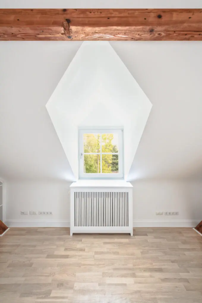 Zentralperspektive einer weißen Dachgaube mit quadratischem Fenster und Heizkörperverkleidung in einem leeren Raum mit Holzboden