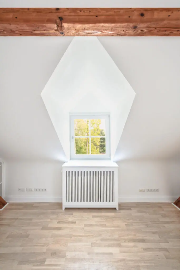 Zentralperspektive einer weißen Dachgaube mit quadratischem Fenster und Heizkörperverkleidung in einem leeren Raum mit Holzboden