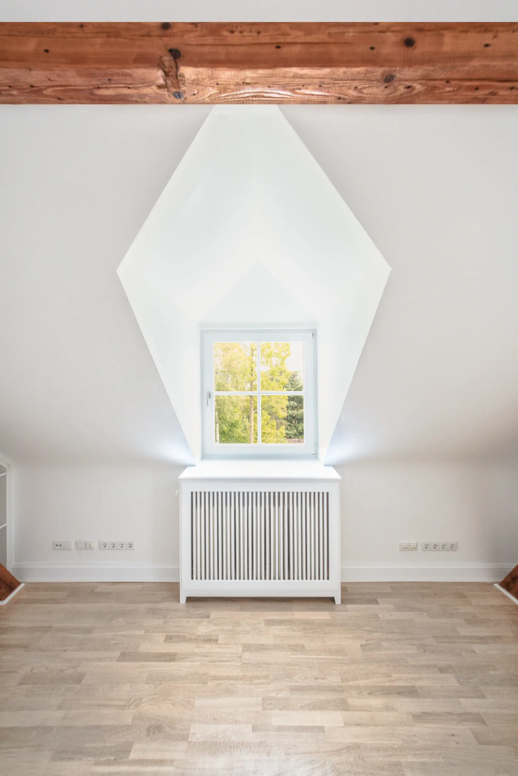 Zentralperspektive einer weißen Dachgaube mit quadratischem Fenster und Heizkörperverkleidung in einem leeren Raum mit Holzboden