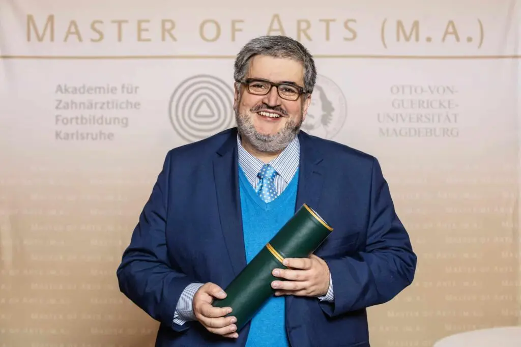 Ein lächelnder Absolvent in formeller Kleidung hält eine grüne Urkundenrolle vor einer akademischen Rückwand mit Schriftzug Master of Arts