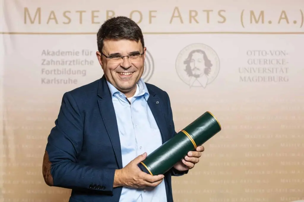 Ein lächelnder Mann in formeller Kleidung hält eine grüne Diplomrolle vor einer Rückwand mit akademischen Logos.