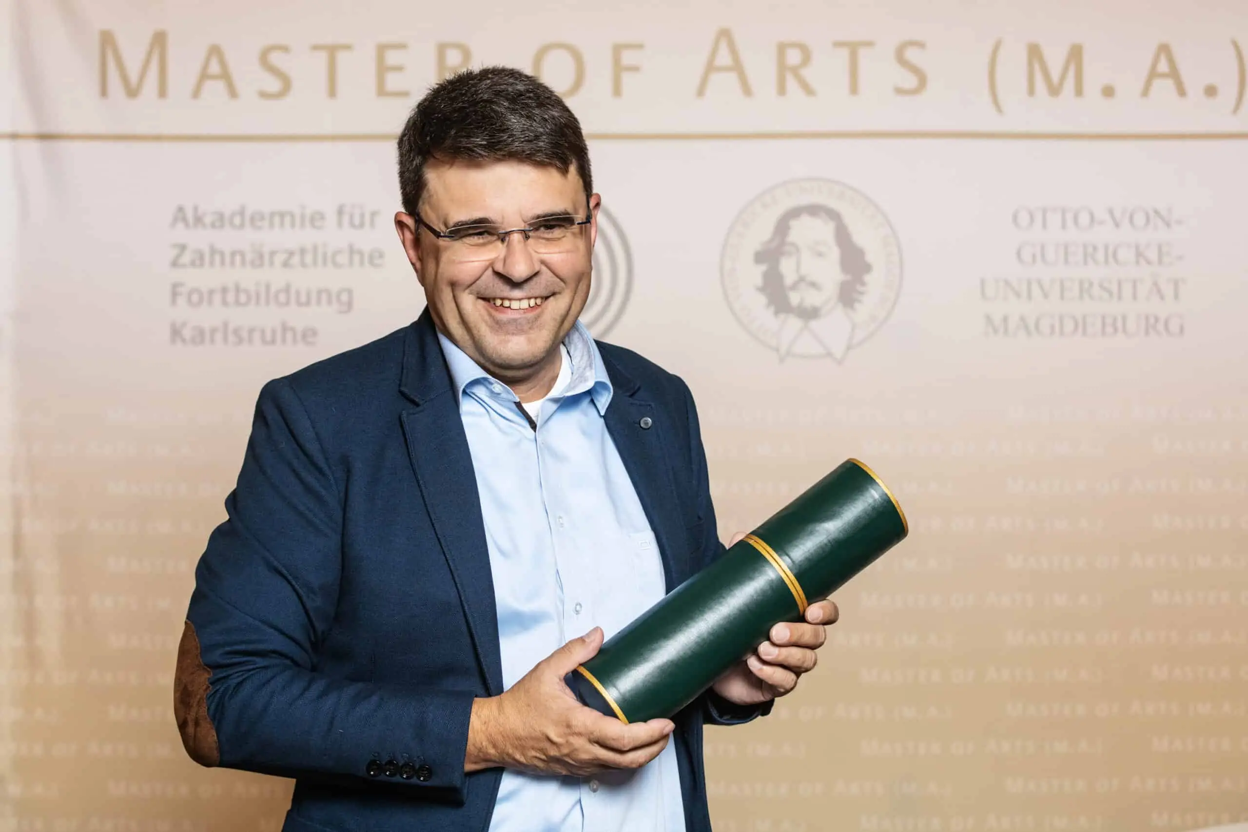 Ein lächelnder Mann in formeller Kleidung hält eine grüne Diplomrolle vor einer Rückwand mit akademischen Logos.