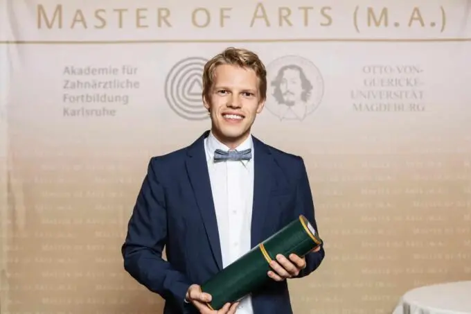 Ein lächelnder Absolvent im Anzug mit Fliege hält eine grüne Zeugnisrolle vor einer Fotowand mit Master of Arts Schriftzug.